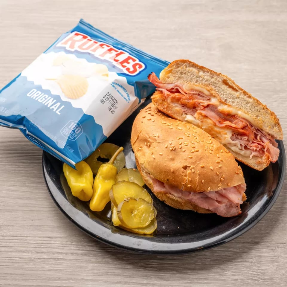 A deli sandwich with ham and cheese, Ruffles potato chips, and pickles on a black plate.