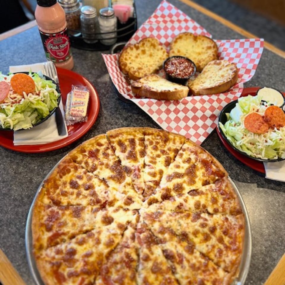 A pizza, garlic bread with marinara, and two side salads with pepperoni and dressing on a table.