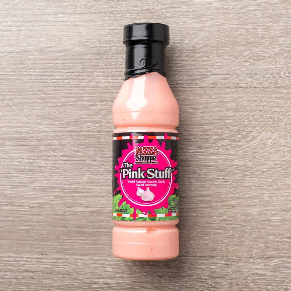 A bottle of The Pizza Shoppe's "Pink Stuff" World-Famous Creamy Garlic Salad Dressing on a wooden surface.
