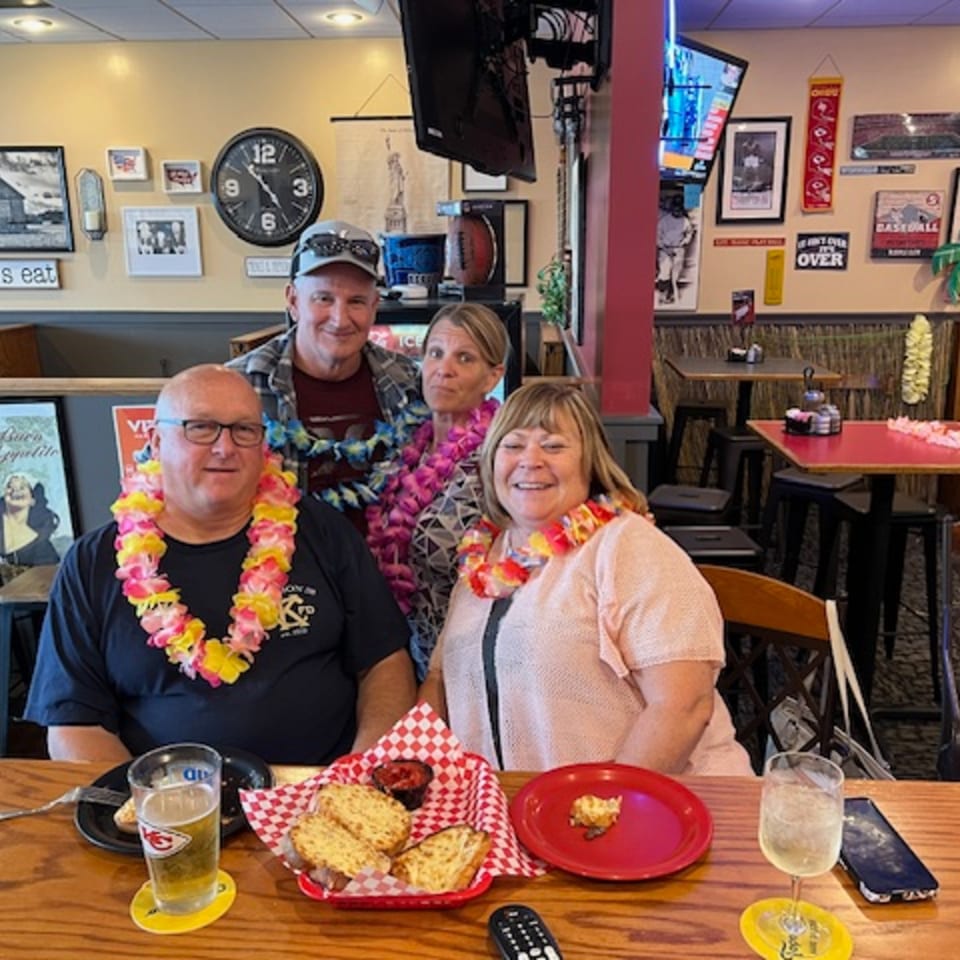 Four smiling people wearing leis sit at a wooden table with food and drinks in a restaurant.