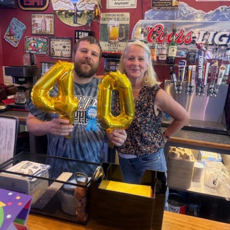 Two people, a man and a woman, holding gold "40" balloons behind a bar with beer taps.