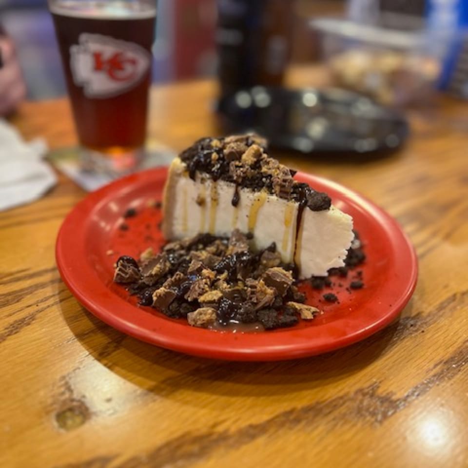 A slice of cheesecake with chocolate and peanut butter toppings on a red plate, with a beer in the background.