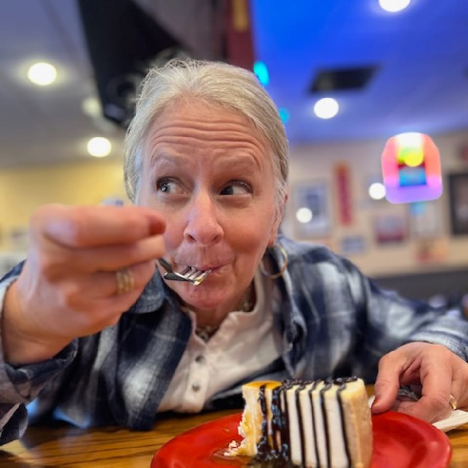 A woman in a plaid shirt enjoys a slice of cheesecake with chocolate drizzle on a red plate.