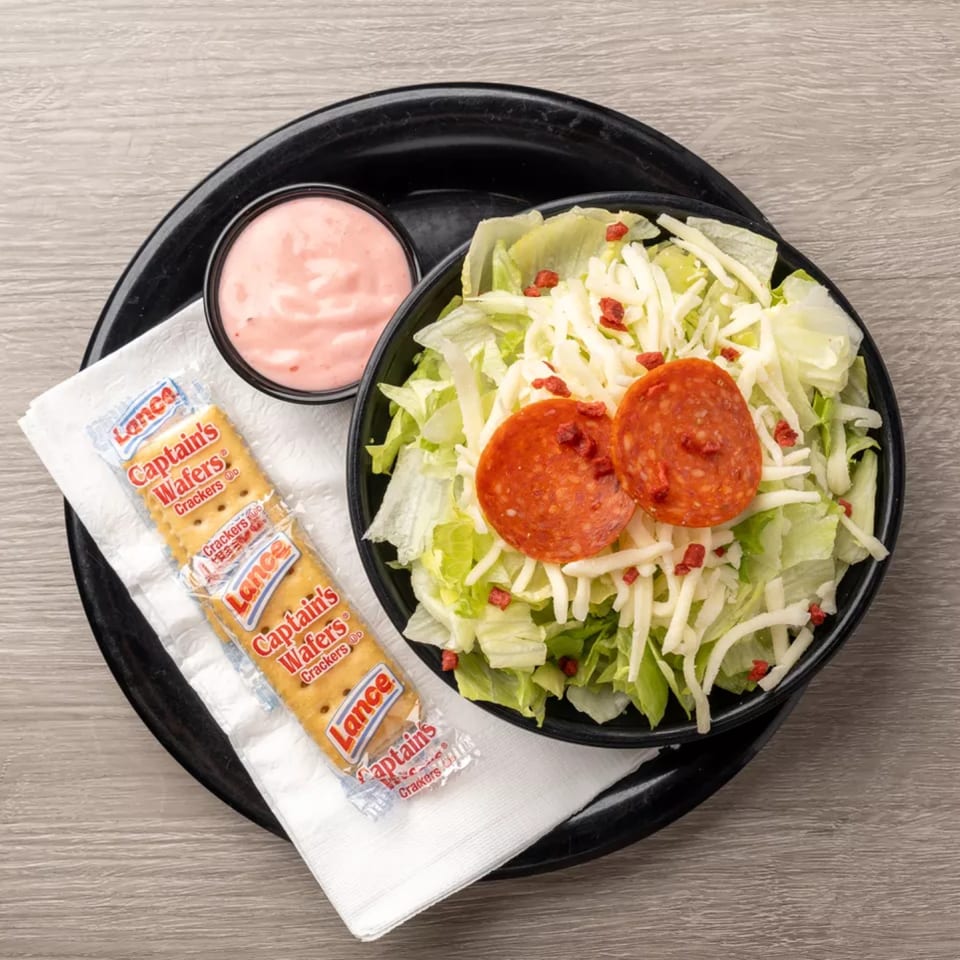 A side salad with pepperoni, shredded cheese, and a packet of Captain's Wafers crackers.