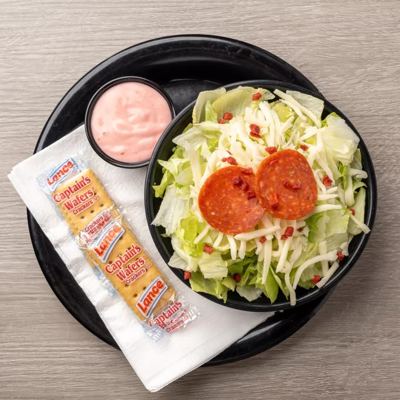 A side salad with pepperoni, shredded cheese, and a packet of Captain's Wafers crackers.
