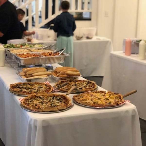 A buffet table with pizzas, hot dog buns, and large trays of food, with people in the background.