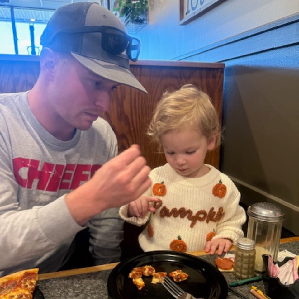 A man in a Chiefs shirt and a child in a pumpkin sweater eating pizza at a restaurant.