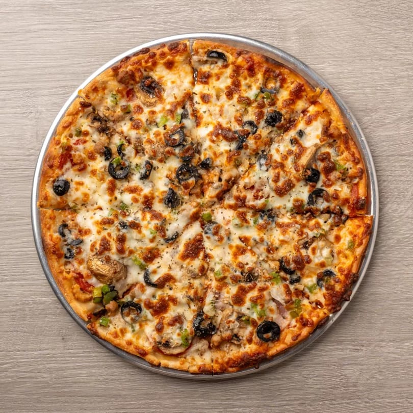 A whole pizza with melted cheese, black olives, mushrooms, and green peppers on a metal pan.