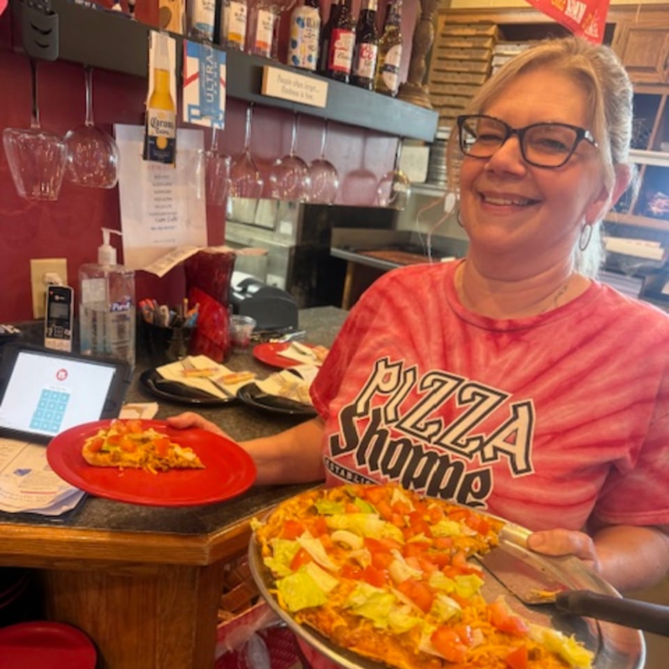 Smiling woman in a Pizza Shoppe shirt holding a pizza with lettuce and tomatoes.