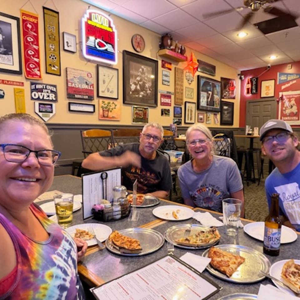 Four smiling people seated at a table with pizza slices and drinks in a casual restaurant.