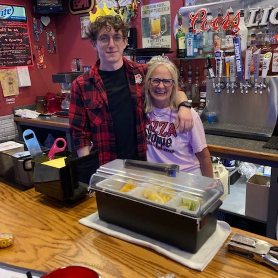 Two people, one in a paper crown, smiling behind a bar with bottles and beer taps.