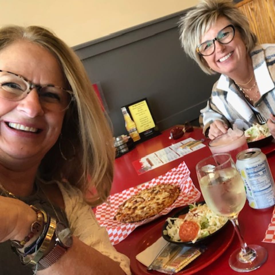Two smiling women enjoying pizza, coleslaw, and drinks at a red table in a restaurant.