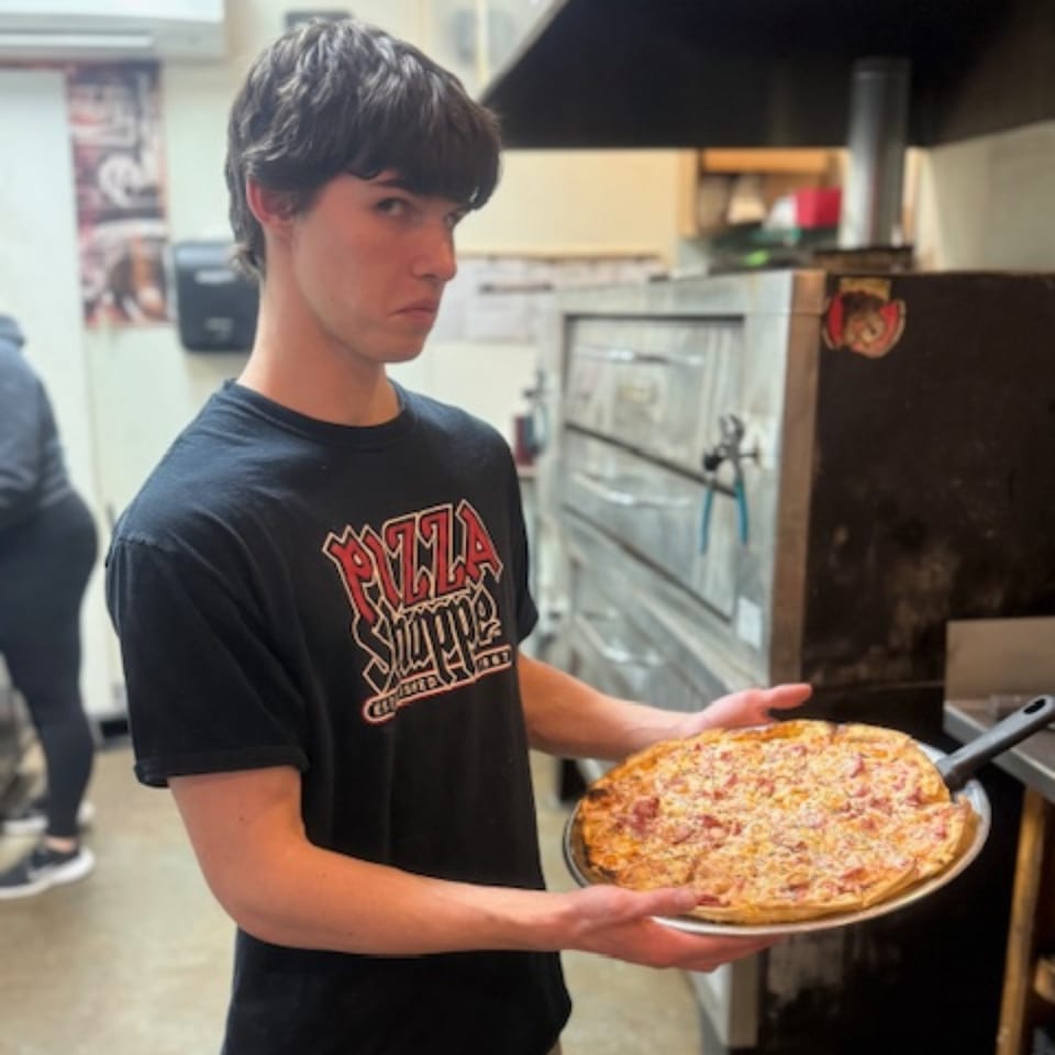 A young man in a "Pizza Shoppe" shirt holds a freshly baked pizza in a kitchen.